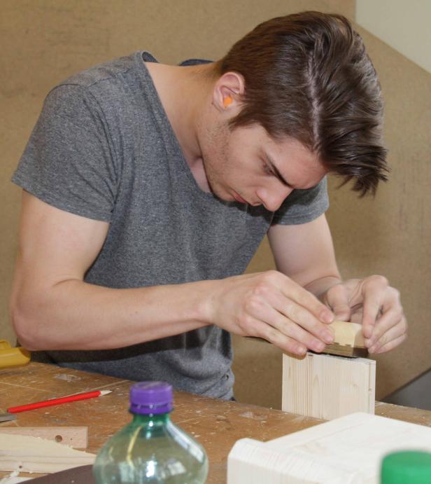 Ein junger Mann hobelt ein Holzstück in einer Werkstatt.