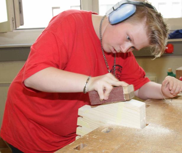Ein Jugendlicher mit Kopfhörern schleift ein Holzstück mit Schleifpapier.