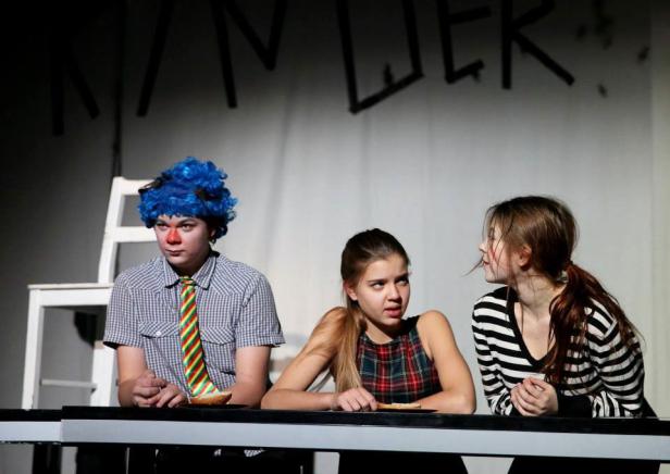 Drei junge Schauspieler sitzen an einem Tisch während einer Theateraufführung.