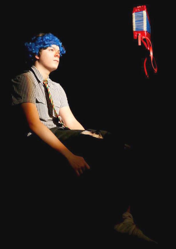 Ein junger Mann mit blauer Perücke sitzt, während ein Akkordeon in der Luft schwebt.