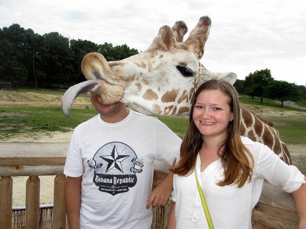 Eine Frau lächelt, während eine Giraffe einem Mann die Zunge ins Gesicht steckt.