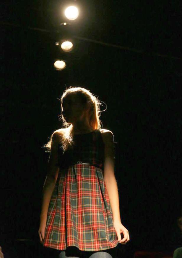 Eine junge Frau mit blonden Haaren und einem karierten Kleid steht im Dunkeln unter Scheinwerfern.