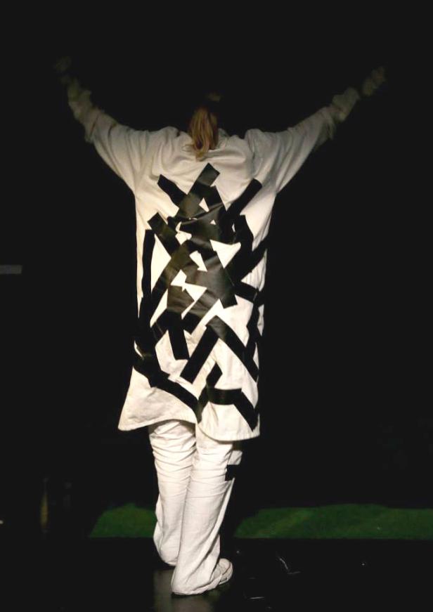 Eine Person in weißer Kleidung mit schwarzem, abstraktem Muster auf dem Rücken steht mit erhobenen Armen da.