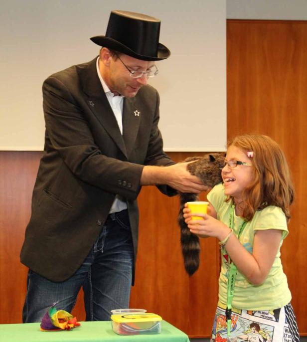 Ein Zauberer mit Zylinder präsentiert einem Mädchen einen Waschbären.