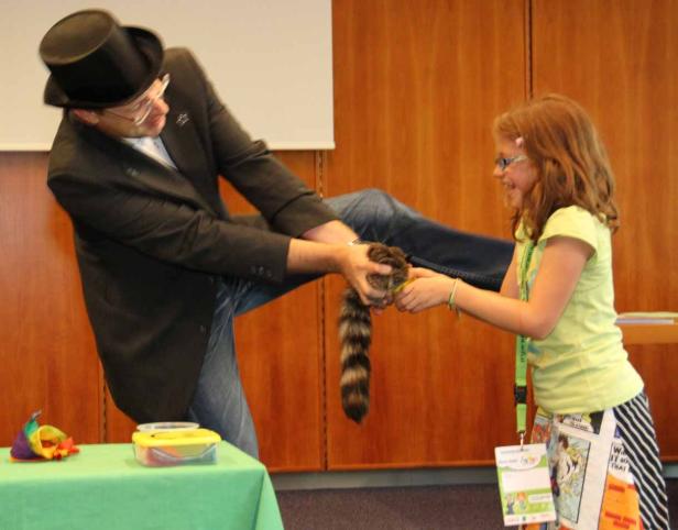 Ein Mann im Anzug und Zylinder übergibt einer lächelnden jungen Frau einen Waschbären.