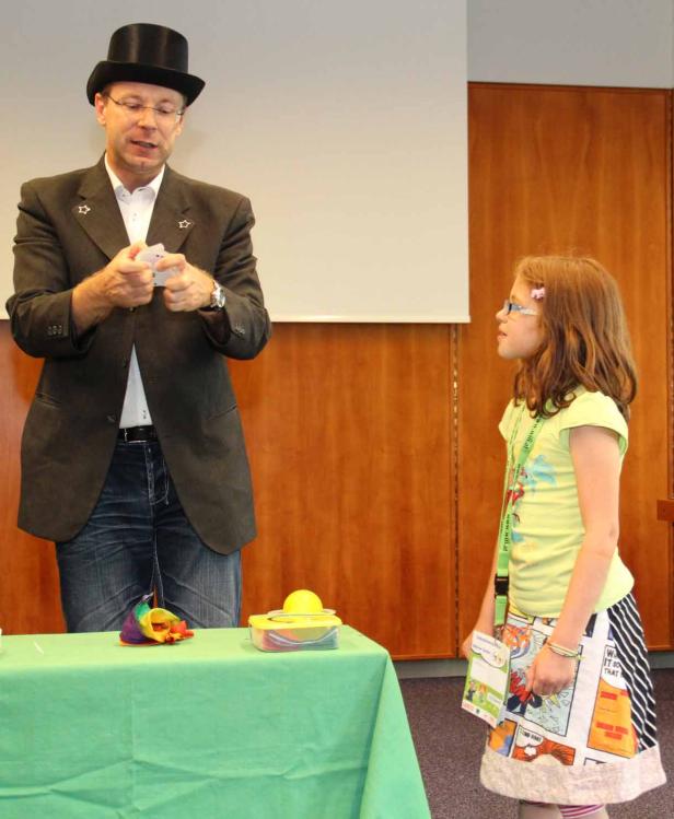 Ein Magier mit Zylinder führt vor einem jungen Mädchen einen Kartentrick vor.