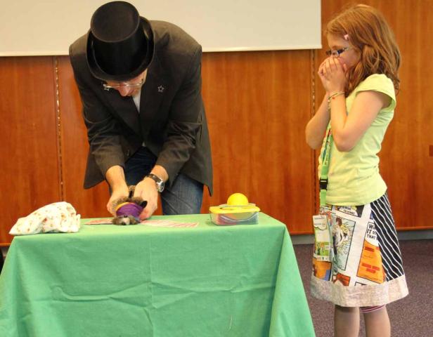 Ein Zauberer mit Zylinder führt einen Trick mit einem kleinen Tier vor, während ein Mädchen zuschaut.