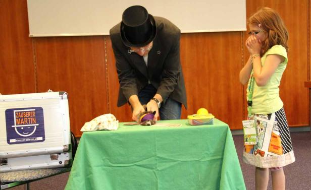 Zauberer Martin führt einen Trick mit einem Tier vor, während ein Mädchen überrascht zusieht.