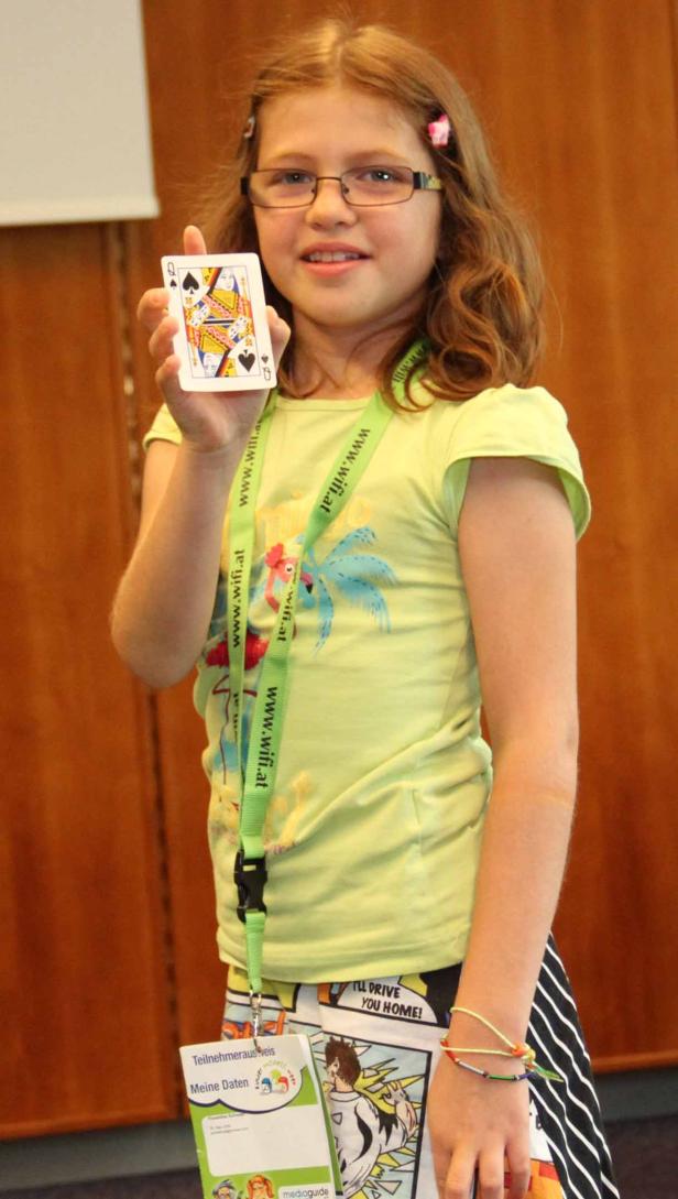 Ein junges Mädchen mit Brille hält eine Spielkarte, die Pik Dame, in der Hand.