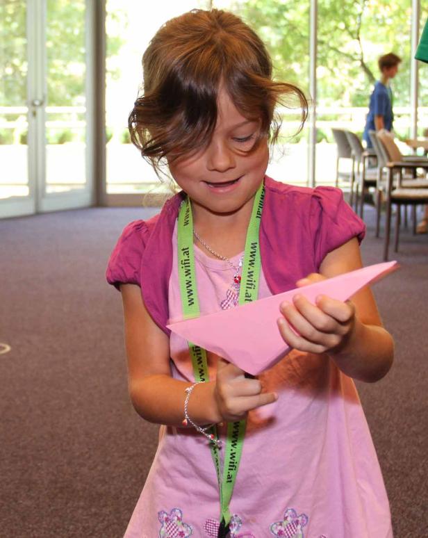 Ein Mädchen faltet konzentriert ein rosafarbenes Papierboot.