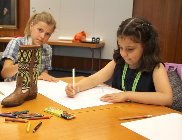Zwei Mädchen sitzen an einem Tisch und zeichnen, ein Cowboy-Stiefel steht daneben.