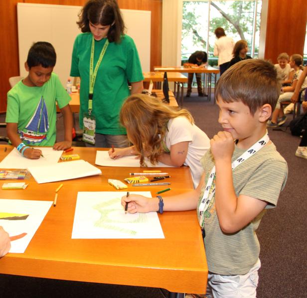 Kinder sitzen an Tischen und malen Bilder mit Stiften und Wachsmalstiften.