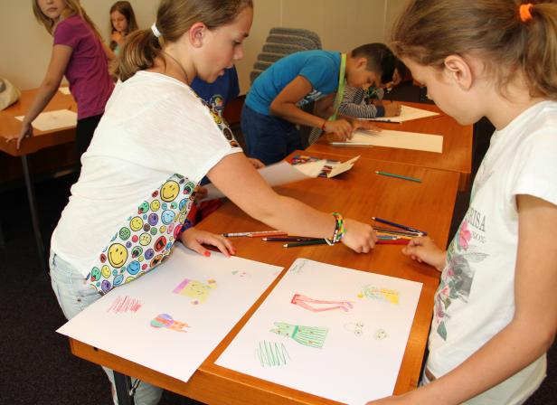 Mehrere Kinder zeichnen Kleider auf Papier an einem langen Tisch.