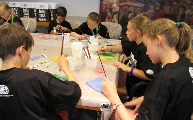 Kinder sitzen an einem Tisch und bemalen Holzfiguren mit Pinseln und Farbe.