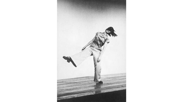 Eine Frau in Hose und Bluse balanciert auf einem Bein.