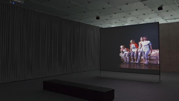 In einem dunklen Raum steht eine Leinwand, auf der eine Gruppe von Personen zu sehen ist.
