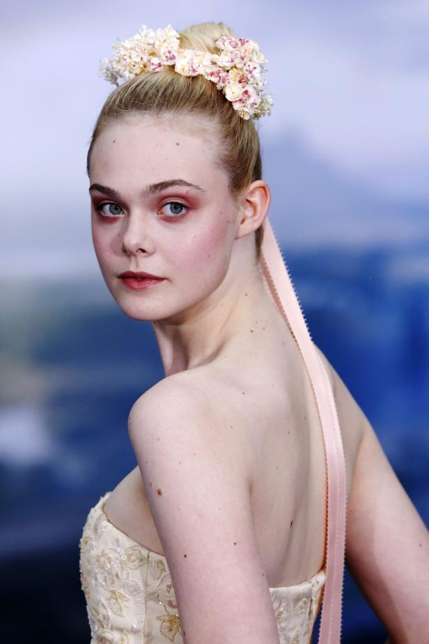 Elle Fanning mit Blumenkranz und hellem Kleid blickt über ihre Schulter.