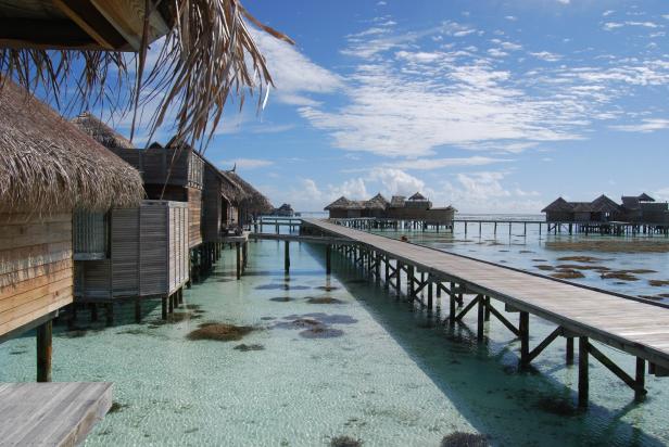 Ein Holzsteg führt zu strohgedeckten Bungalows über dem türkisfarbenen Wasser.