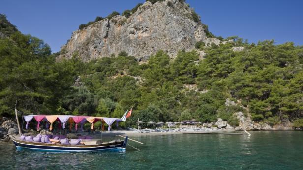 Ein Boot mit Baldachin liegt vor einem Strand mit Liegestühlen und einem bewaldeten Felsen im Hintergrund.