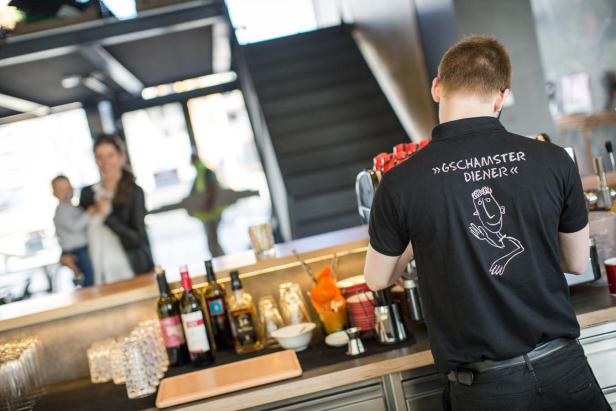 Ein Barkeeper mit einem T-Shirt mit der Aufschrift „Gschamster Diener“ arbeitet hinter der Bar.
