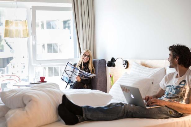 Ein Paar entspannt im Bett; sie liest Zeitung, er arbeitet am Laptop.