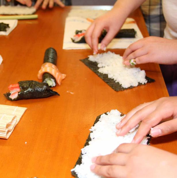 Mehrere Personen bereiten Sushi zu, indem sie Reis auf Noriblätter legen.