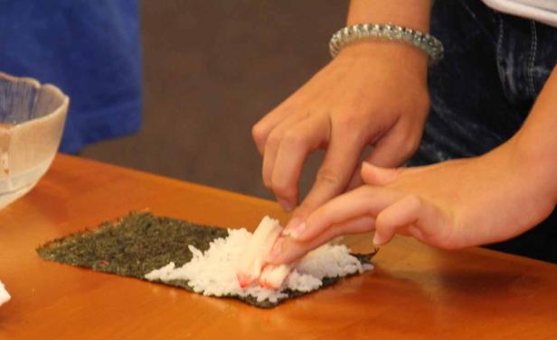 Eine Person bereitet Sushi mit Reis und Krebsfleisch auf einem Noriblatt zu.