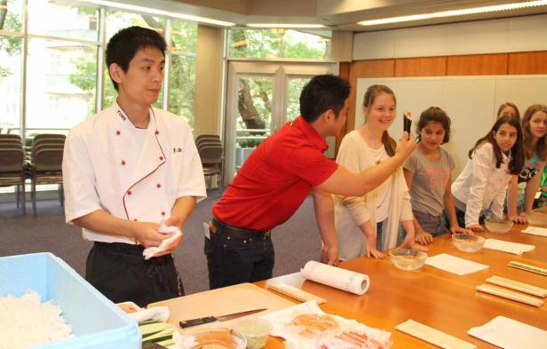 Ein Koch und ein Mann im roten Hemd leiten einen Sushi-Kurs für eine Gruppe junger Mädchen.