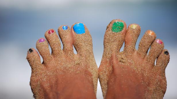 Zwei sandbedeckte Füße mit bunten Zehennägeln vor einem verschwommenen Hintergrund.