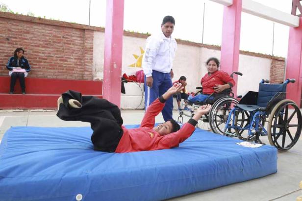 Ein Junge liegt auf einer blauen Matte, während andere Personen zusehen, darunter eine Frau im Rollstuhl.