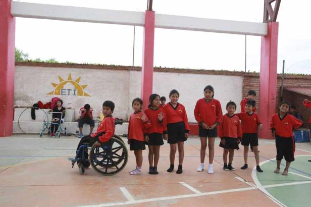 Eine Gruppe von Schulkindern in roten Uniformen steht auf einem Sportplatz, ein Junge sitzt im Rollstuhl.