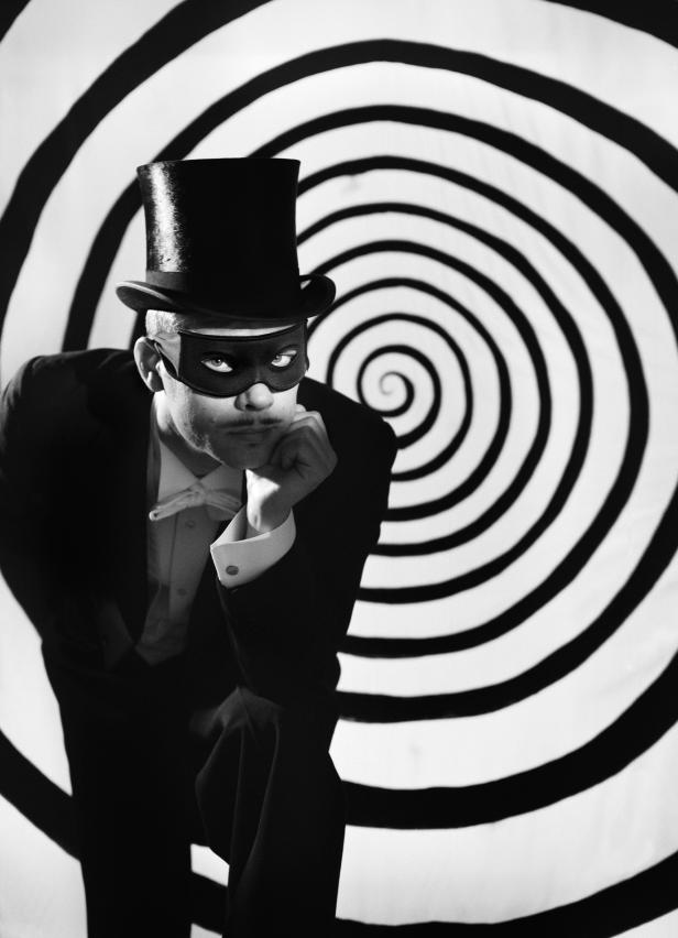 Ein Mann mit Zylinder und Maske posiert vor einem hypnotischen Hintergrund.
