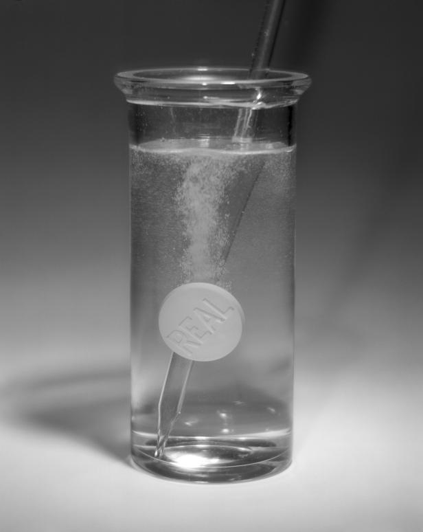 Eine Brausetablette mit der Aufschrift „REAL“ löst sich in einem Glas Wasser auf.