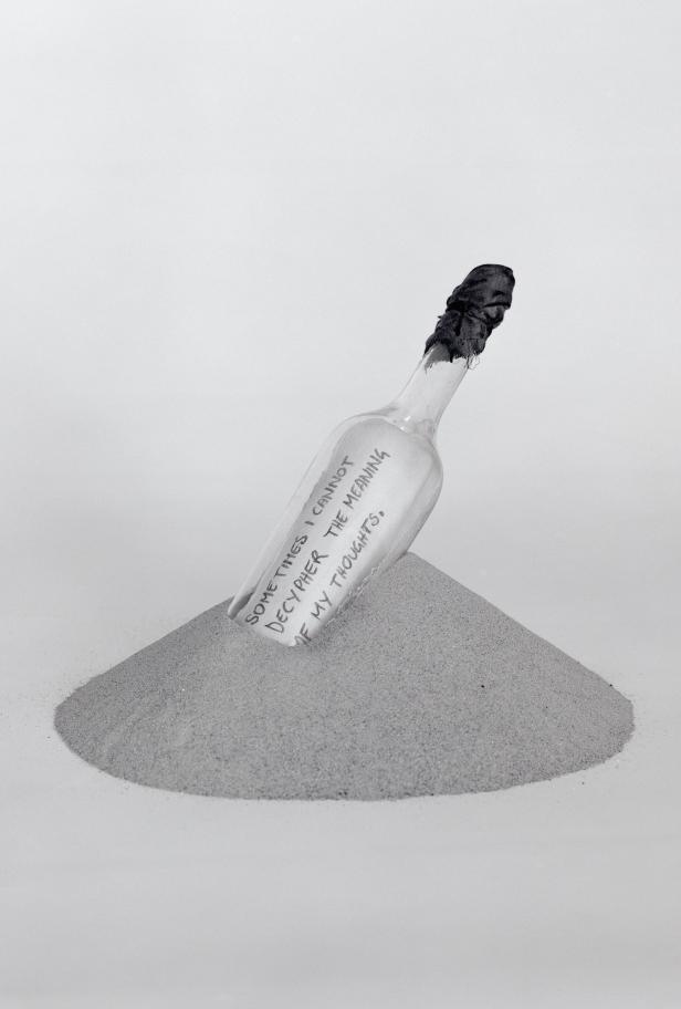 Eine Glasflasche mit der Aufschrift „Sometimes I cannot decipher the meaning of my thoughts“ steckt in einem Sandhaufen.
