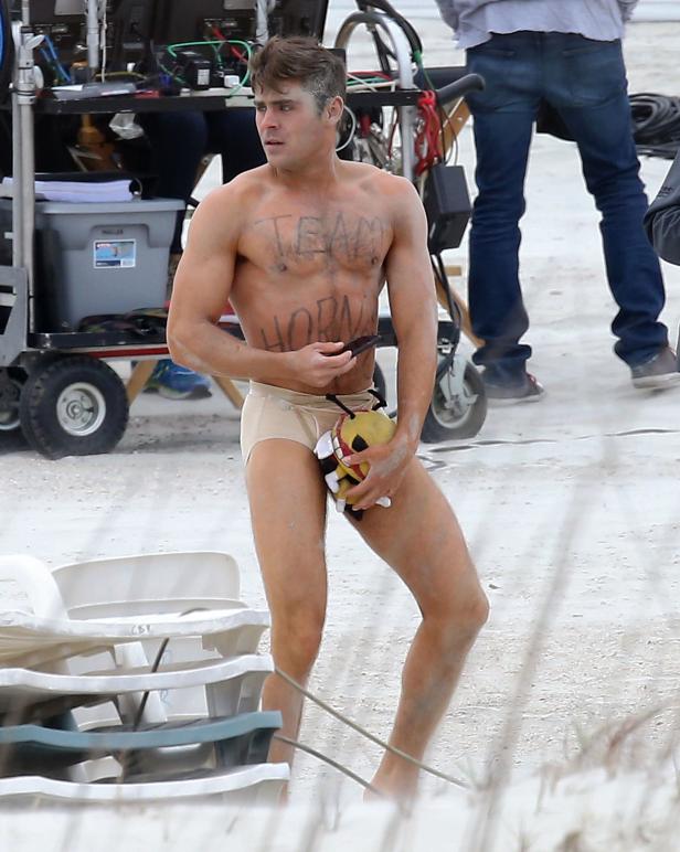 Zac Efron am Strand mit aufgemaltem „Team Horny“-Schriftzug auf der Brust und einer Plüschbiene in der Hand.