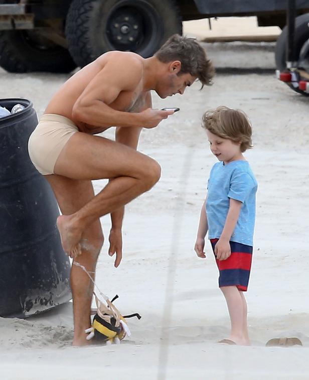 Zac Efron steht am Strand und schaut auf sein Handy, während ein kleiner Junge ihn ansieht.