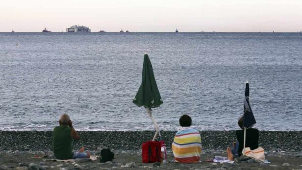 Menschen sitzen am Strand unter Sonnenschirmen und blicken auf das Meer mit Schiffen am Horizont.