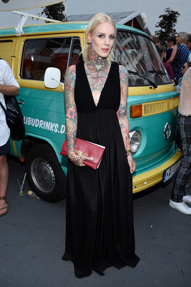 Eine tätowierte Frau in einem schwarzen Kleid posiert vor einem gelb-türkisen VW-Bus.