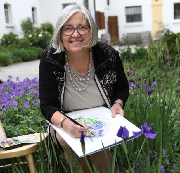 Eine ältere Frau mit Brille sitzt im Garten und malt ein Bild von blauen Irisblüten.