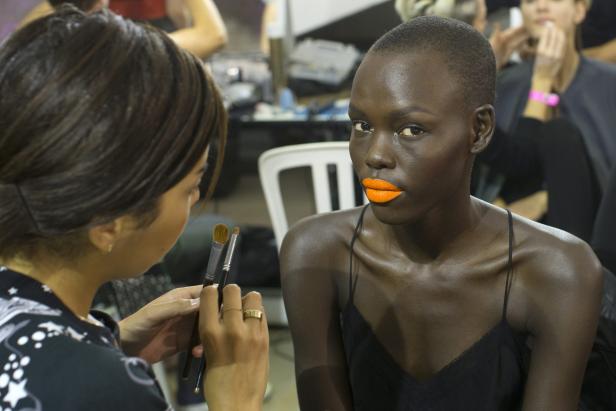 Eine Frau mit orangefarbenem Lippenstift wird von einer Maskenbildnerin geschminkt.