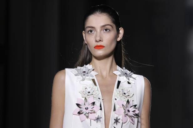 Eine Frau mit orangefarbenem Lippenstift trägt ein weißes Kleid mit Blumenapplikationen.