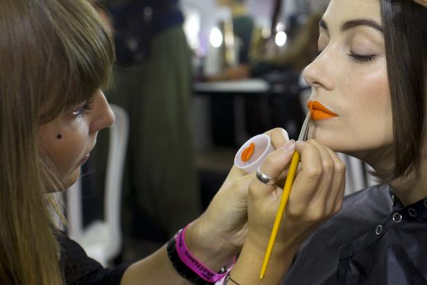Eine Visagistin trägt orangefarbenen Lippenstift auf das Gesicht eines Models auf.