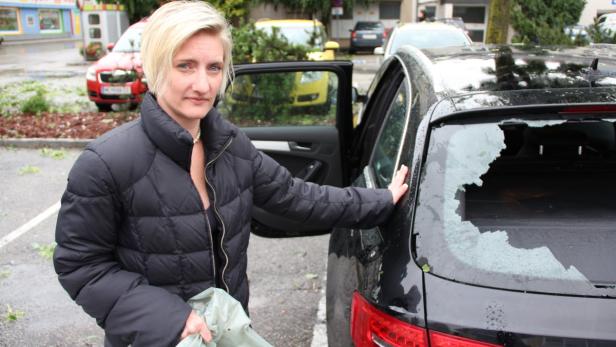Eine Frau steht neben einem Auto mit einer durch Hagel beschädigten Heckscheibe.