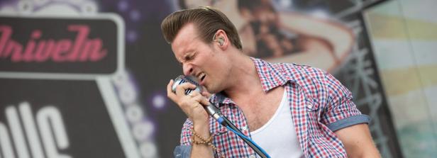 Ein Mann mit Pompadour-Frisur singt in ein Mikrofon vor einer Bühne.