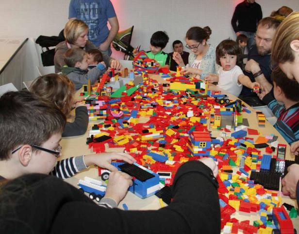 Kinder und Erwachsene bauen gemeinsam mit bunten Legosteinen an einem Tisch.