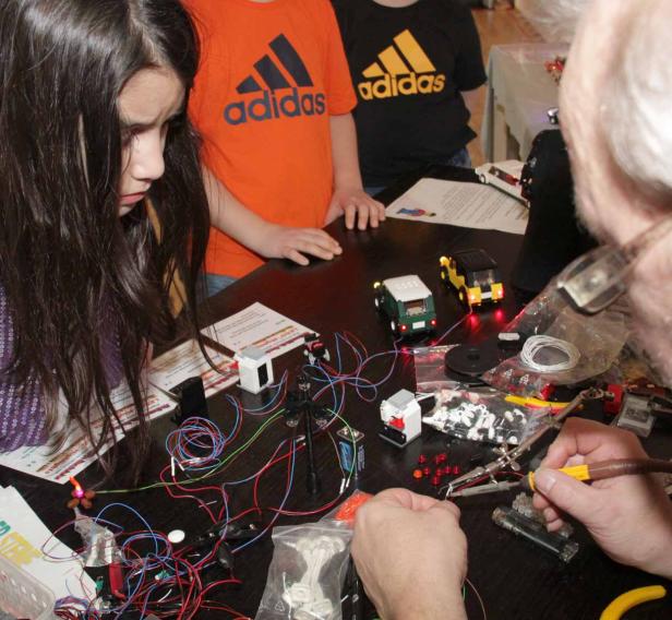 Ein Mann lötet an einer Platine, während Kinder Lego-Modelle mit Lichtern betrachten.