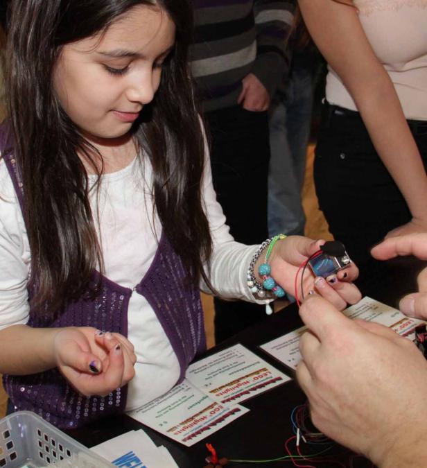 Ein junges Mädchen hantiert mit einer Batterie und Kabeln an einem Tisch mit LEGO-Materialien.