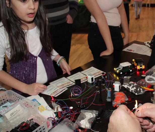 Ein Mädchen betrachtet einen Tisch mit Lego-Bauteilen und elektronischen Komponenten, während jemand einen Lötkolben benutzt.