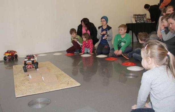 Kinder und Erwachsene beobachten ein ferngesteuertes Auto auf einer Rampe.