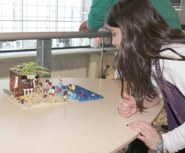 Ein Mädchen betrachtet ein Lego-Diorama einer Strandszene mit Figuren und einem Kiosk.